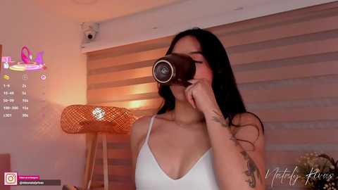 Video of an Asian woman with long black hair, wearing a white tank top, taking a selfie with a camera in front of her face. Background includes a calendar, a wicker lamp, and a striped blind.