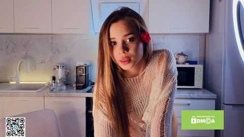 Media: Video of a young woman with long, straight brown hair adorned with a red flower, wearing a beige knit sweater, leaning forward in a modern kitchen with white cabinets, marble backsplash, and a microwave.