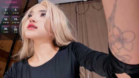 Media: Video of a blonde woman with a black top, red lipstick, and a tattoo on her arm, standing in a dimly lit room with a calendar display in the background.