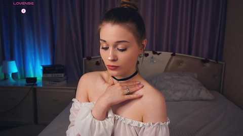 Media: A video of a young woman with light skin and brown hair in a bun, wearing a black choker and white off-shoulder top, standing in a dimly-lit bedroom with purple curtains.