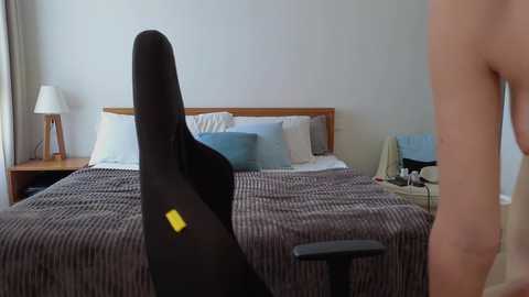 Media: Video of a modern bedroom with a wooden bed, beige sheets, a black gaming chair, a wooden side table with a lamp, and a person in the background.