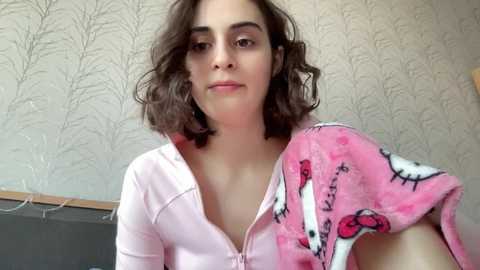 Media: Video of a young woman with wavy brown hair, wearing a pink hoodie and a pink blanket with Hello Kitty design. Background features a beige, textured wallpaper.