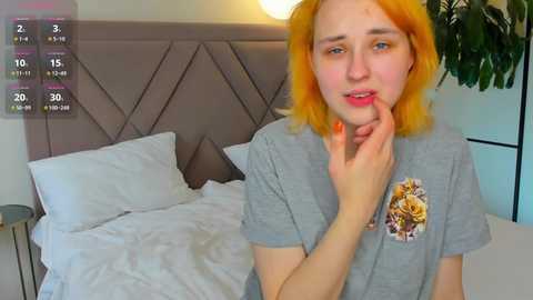 Media: Video of a young woman with shoulder-length, bright orange hair, fair skin, wearing a gray T-shirt, standing near an unmade bed in a modern bedroom.