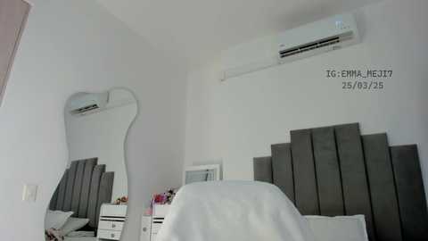 Media: Video of a minimalist bedroom with a gray upholstered headboard, a white bedspread, a mirror, and an air conditioner on the wall.