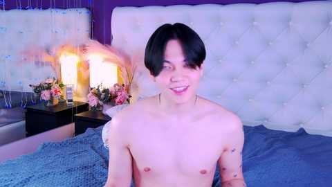 Media: A video of a shirtless, young, slim Asian man with short black hair, small breasts, and a tattoo on his left arm, smiling in a modern bedroom with a tufted headboard, floral decor, and blue bedding.