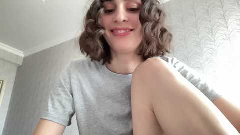 Media: A young woman with curly brown hair and light skin, smiling, wearing a gray t-shirt, sitting in a room with a gray wallpaper and white trim.