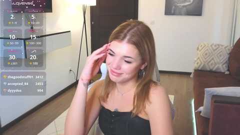 Media: Video of a young Caucasian woman with light skin and straight, shoulder-length blonde hair, wearing a black strapless top, sitting in a modern living room.