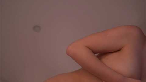 Media: A video of a nude, light-skinned person with medium-sized breasts, partially obscured by their arm, against a plain, beige wall. The image is soft-focused, with a subtle, warm lighting tone.