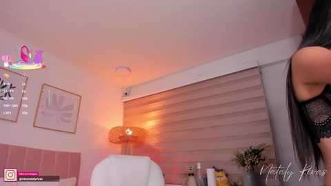 A video of a cozy, well-lit room with a woman's bare arm visible, pink walls, a lamp, and a partially open window with blinds.
