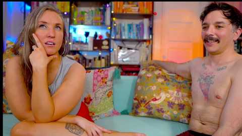 Media: Video of a topless man with a mustache, tattooed, and a woman in a gray shirt, sitting on a colorful couch in a cluttered room with shelves of items in the background.