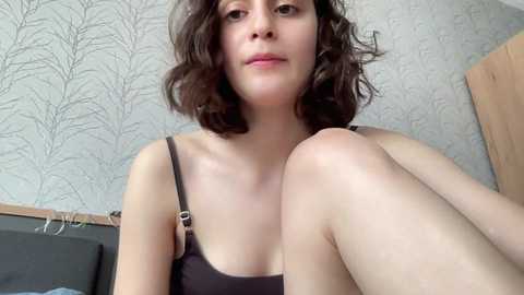 Media: A video of a fair-skinned woman with shoulder-length, wavy brown hair, wearing a black tank top, sitting on a bed with a wooden headboard and gray-patterned wallpaper.