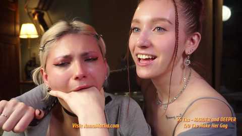 Media: Video of two young women, one with blonde hair in a bun, the other with brown hair, smiling, in a dimly lit room.