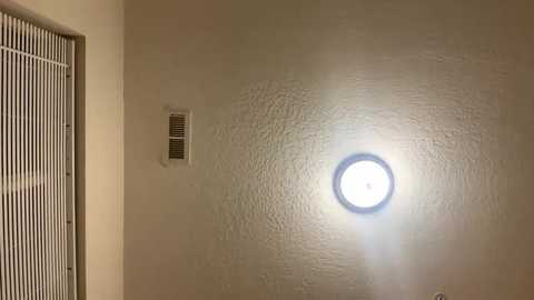 Media: Video of a beige textured ceiling with a circular, bright light fixture emitting a soft glow. The ceiling is adjacent to a white window with vertical blinds, creating a warm, utilitarian interior setting.