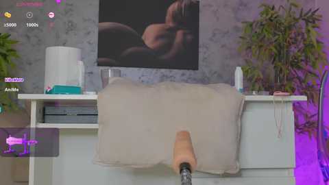 A video of a person with a realistic, tan, dildo-like object being inserted into a beige cushioned chair, set in a modern, dimly lit room with plants and a blurred, abstract art piece on the wall.