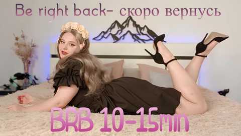 Media: A video of a plus-sized woman with fair skin, long brown hair, and a black dress lying provocatively on a bed. Text reads \"Be right back- 10-15min\" in Russian.