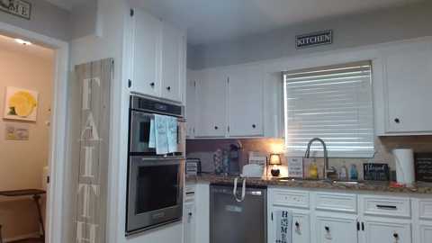 Media: Video of a modern kitchen with white cabinets, granite countertops, stainless steel appliances, and a window with closed blinds. The room is clean and organized with a \"Kitchen\" sign above the cabinets.