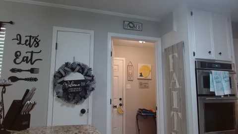 Media: Video of a small kitchen with white cabinets, black door, and \"Let's eat\" sign. A black and white wreath hangs on the door. Microwave, oven, and utensils are visible.