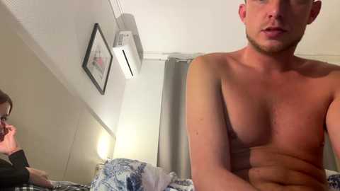 Media: Video of a shirtless, muscular man with short hair, sitting on a bed in a dimly lit, beige room.