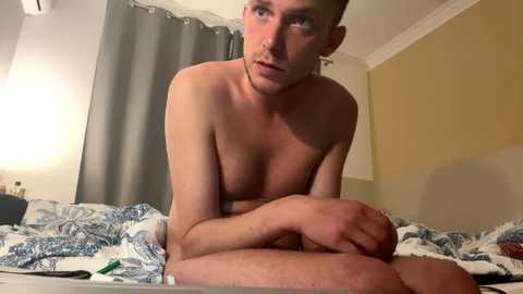 Media: Video of a shirtless, light-skinned, young man with short hair, sitting on a bed with blue and white patterned sheets. He has a serious expression, and the room has beige walls, a grey curtain, and a bedside lamp.