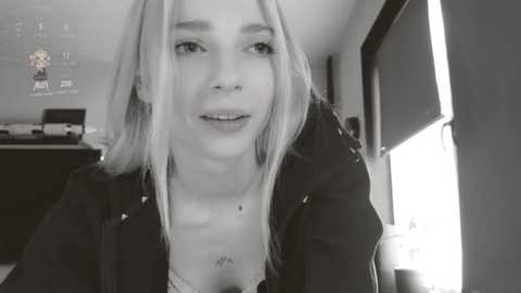 Media: A black-and-white video of a young woman with long, straight hair, wearing a black jacket, seated near a window in a dimly lit room.