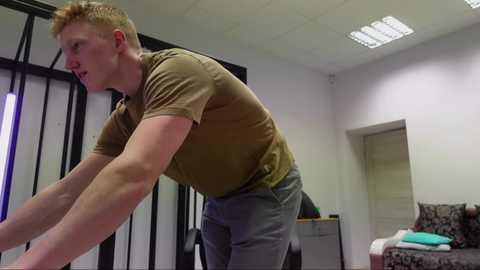 Media: Video of a tall, muscular, fair-skinned man with short red hair, wearing a brown t-shirt and gray pants, leaning forward, in a modern, sparsely furnished room with white walls and a fluorescent ceiling light.