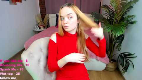 Video of a young woman with long blonde hair, wearing a red turtleneck, in a cozy bedroom with a white bed, plants, and pink throw pillows.
