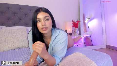 Media: Video of a Latina woman with long black hair and light skin, wearing a light blue shirt, lying on a bed in a modern bedroom with grey headboard, white walls, and a purple lamp.