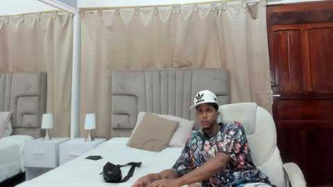 Media: Video of a man in a floral shirt, white bucket hat, sitting on a bed with beige headboard, beige curtains, wooden door, and bedside table lamps.