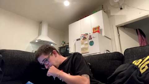 Media: Video of a young man with glasses and short brown hair, lying on a dark gray couch, wearing a black T-shirt, in a cozy, modern living room with white cabinets and a refrigerator.