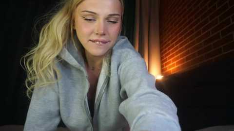 Media: A video of a blonde woman with fair skin, wearing a gray hoodie, taking a selfie in a dimly lit room with a brick wall and a warm light source.