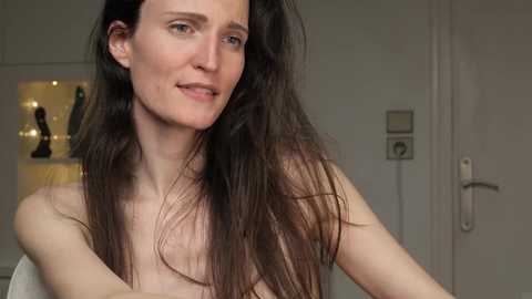 Media: Video of a fair-skinned woman with long, dark brown hair, smiling slightly, topless, standing in a dimly lit room with a white door and a shelf holding a black dildo.