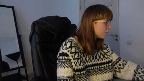 Media: Video of a young woman with shoulder-length auburn hair, wearing glasses, a black and white geometric-patterned sweater, and sitting indoors. Background features a black leather chair, a mirror, and a white door.