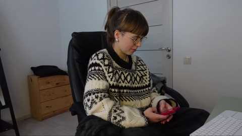 Media: Video of a young woman with glasses, brown hair tied in a ponytail, wearing a black and white patterned sweater, sitting on a black office chair in a minimalist office space with a white door and wooden drawers.