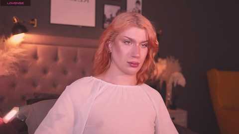 Media: Video of a fair-skinned woman with wavy, shoulder-length, orange hair, wearing a white blouse, seated indoors with a dark grey, tufted headboard and decorative pillows in the background.