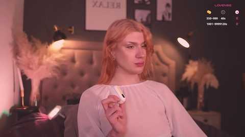 Media: Video of a pale-skinned, red-haired woman with delicate features, wearing a sheer white blouse, holding a vape pen. Background features a dark room with modern decor, including a tufted headboard, black lamp, and feather decor.