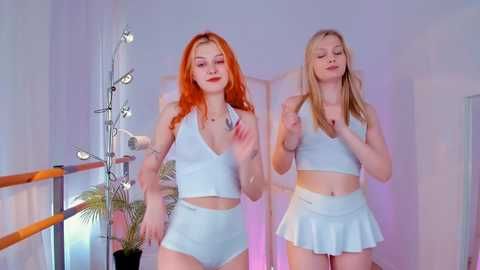 Media: Video of two young women with vibrant red and blonde hair, wearing matching light blue crop tops and short skirts, standing against a minimalist backdrop with a plant and a lamp.
