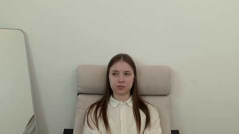 Video of a young Caucasian woman with long, straight brown hair, wearing a white blouse, sitting in a beige office chair against a plain white wall, with a large mirror to the left.
