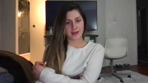 Video of a young woman with long brown hair, wearing a white long-sleeved top, sitting on a bed in a modern, dimly-lit bedroom with a TV, chair, and open door.