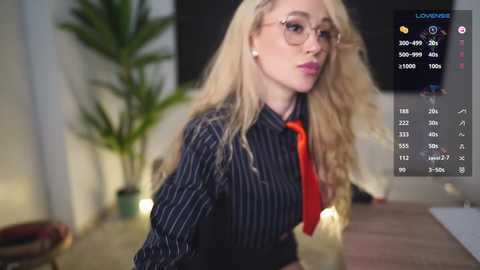 Media: Video of a blonde woman with glasses in a dark pinstripe shirt and red tie, leaning slightly forward, in a modern indoor setting with a potted plant and a digital display showing statistics.