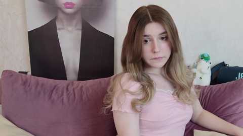 Media: Video of a young Caucasian woman with long, wavy blonde hair, wearing a pink short-sleeved top, sitting on a mauve couch. Behind her, a large black-and-white portrait of a woman with pink lips is displayed on a white wall.