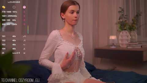 A video captures a young woman with light skin and brown hair, wearing a white crochet top and skirt, seated on a bed in a dimly lit bedroom.