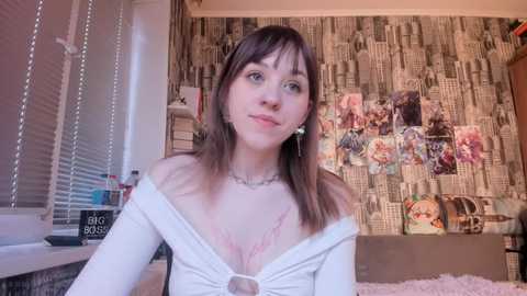 Video of a young woman with light skin and straight brown hair, wearing a white off-shoulder top, sitting in a room with cityscape wallpaper, a bed with pink blanket, and framed photos.