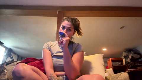 Video of a young woman with light brown skin, wearing a gray t-shirt, sitting cross-legged on a bed with a red blanket, holding a blue phone to her mouth. Background shows a wooden ceiling and a furry dog.