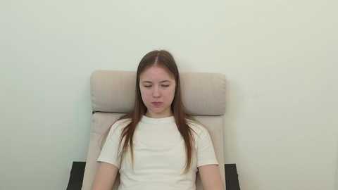 Video of a Caucasian woman with straight, brown hair, wearing a white T-shirt, seated in a beige armchair against a plain white wall, appearing relaxed and contemplative.