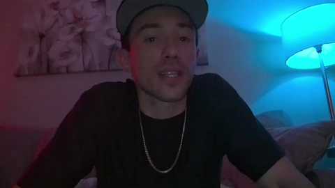 Media: Video of a young, light-skinned man with a trimmed beard, wearing a grey cap, black t-shirt, and silver necklace, smiling. Background features a floral painting and a blue-lit lamp.