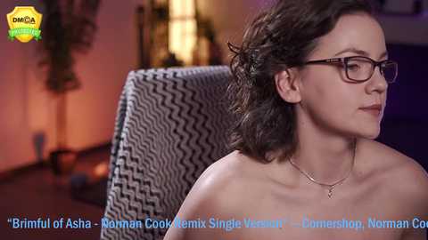 Video of a fair-skinned, curly-haired woman with glasses, wearing a necklace, in a dimly lit room with a black and white patterned chair.