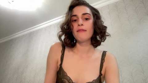 Media: Video of a young woman with wavy, shoulder-length brown hair, fair skin, and light makeup. She wears a black lace bra, standing against a textured, light-colored wallpaper. The room has a modern, minimalist aesthetic.