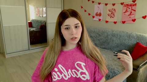 Media: Video of a young woman with long, straight brown hair, wearing a pink \"Barbie\" t-shirt, standing in a modern, beige-walled bedroom with a gray couch, heart decorations, and a mirrored wardrobe.