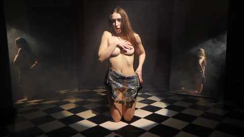 Media: Video of a topless, fair-skinned, brunette woman kneeling on a black-and-white checkered floor, covering her breasts with one hand, wearing a short, patterned skirt, in a dimly lit, mirrored room with a shadowy figure in the background.