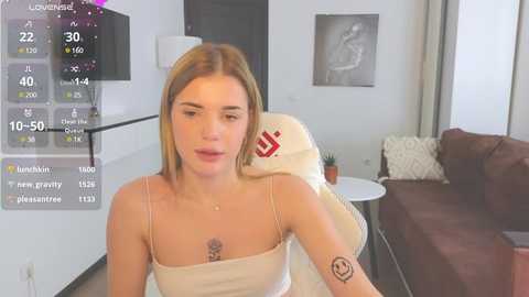 Media: A video of a young Caucasian woman with straight, shoulder-length blonde hair, wearing a beige camisole, sitting in a modern living room with a beige armchair, a brown sofa, and a white wall.
